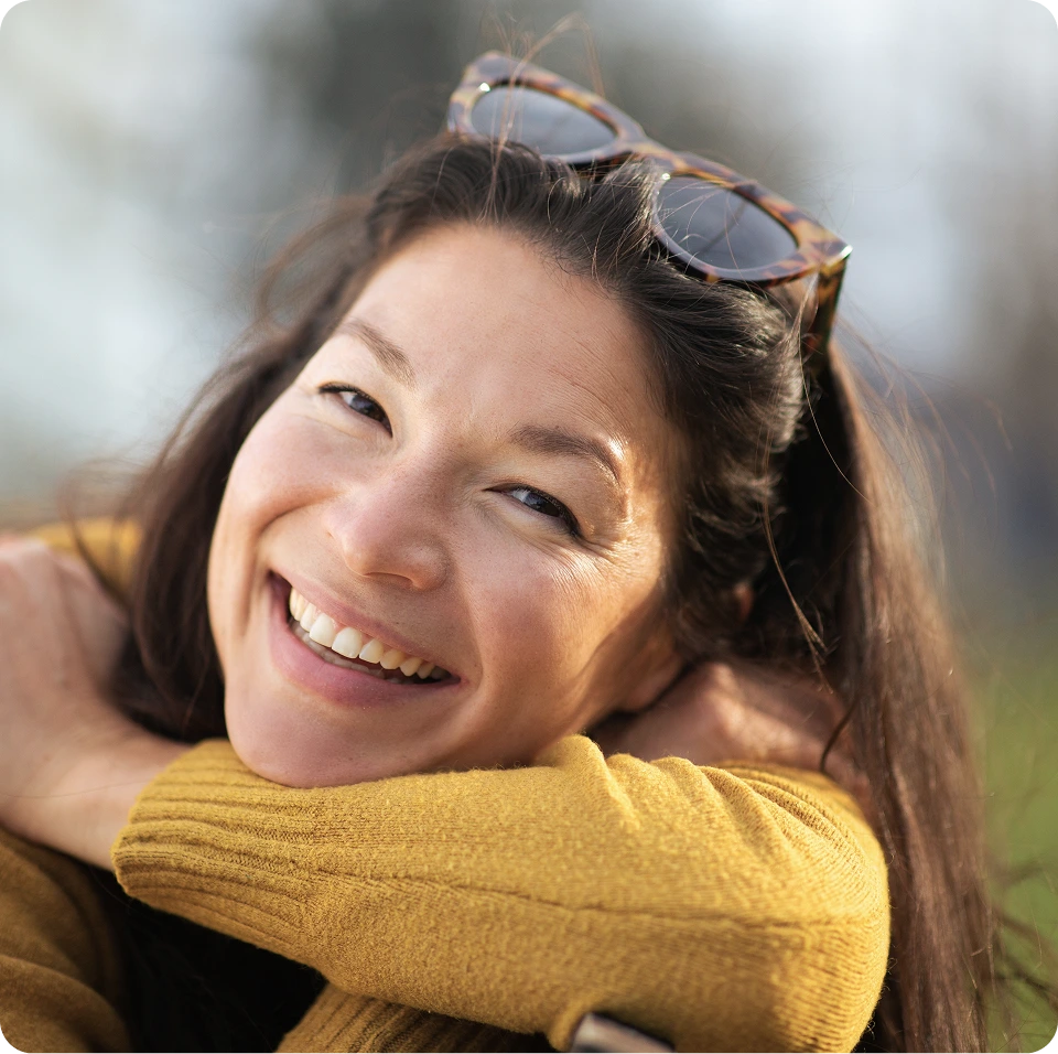 Woman with sunglasses on head wearing yellow sweater smiling with blurred background