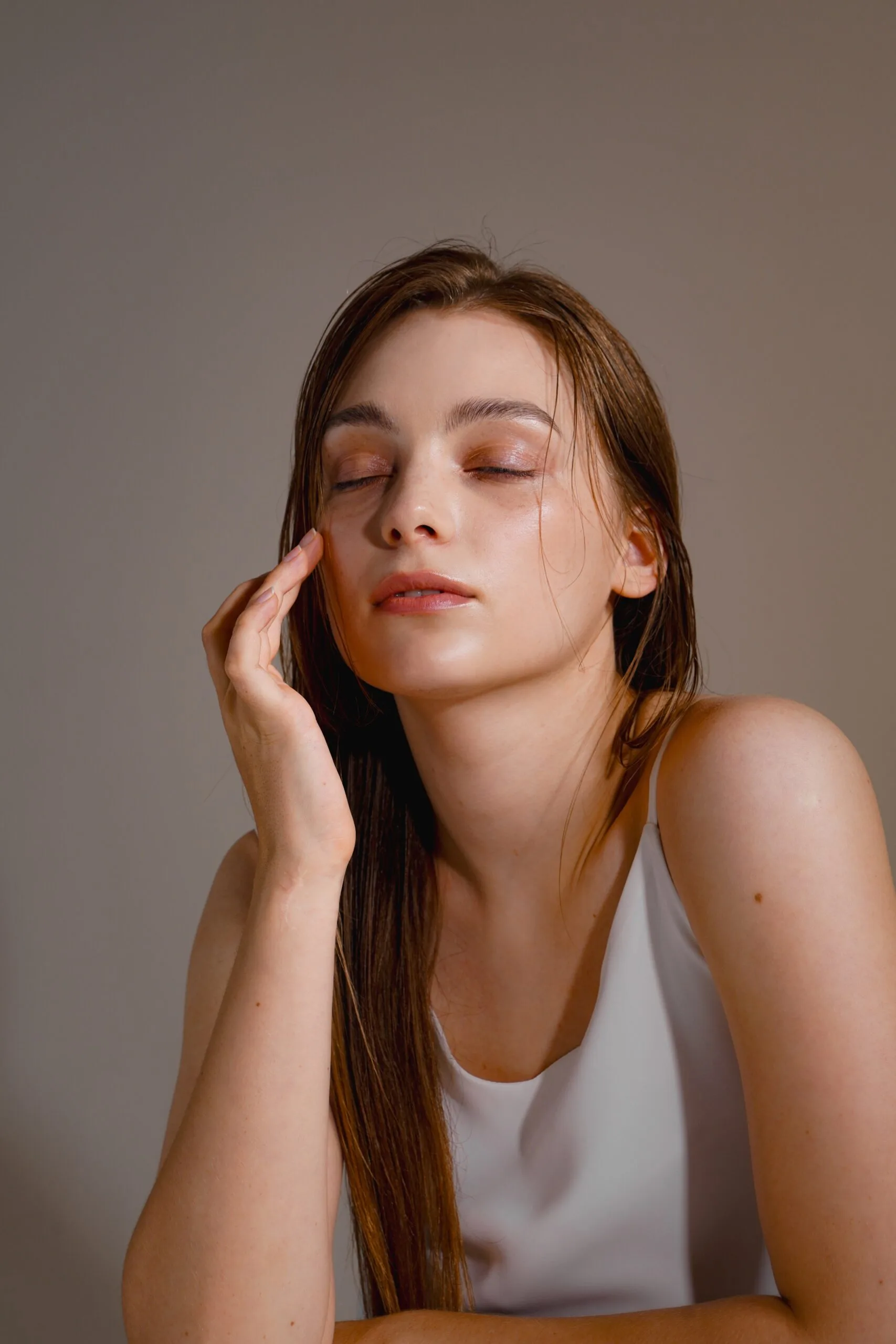 Woman with glowing, smooth skin touching her cheek and looking peaceful against a neutral background