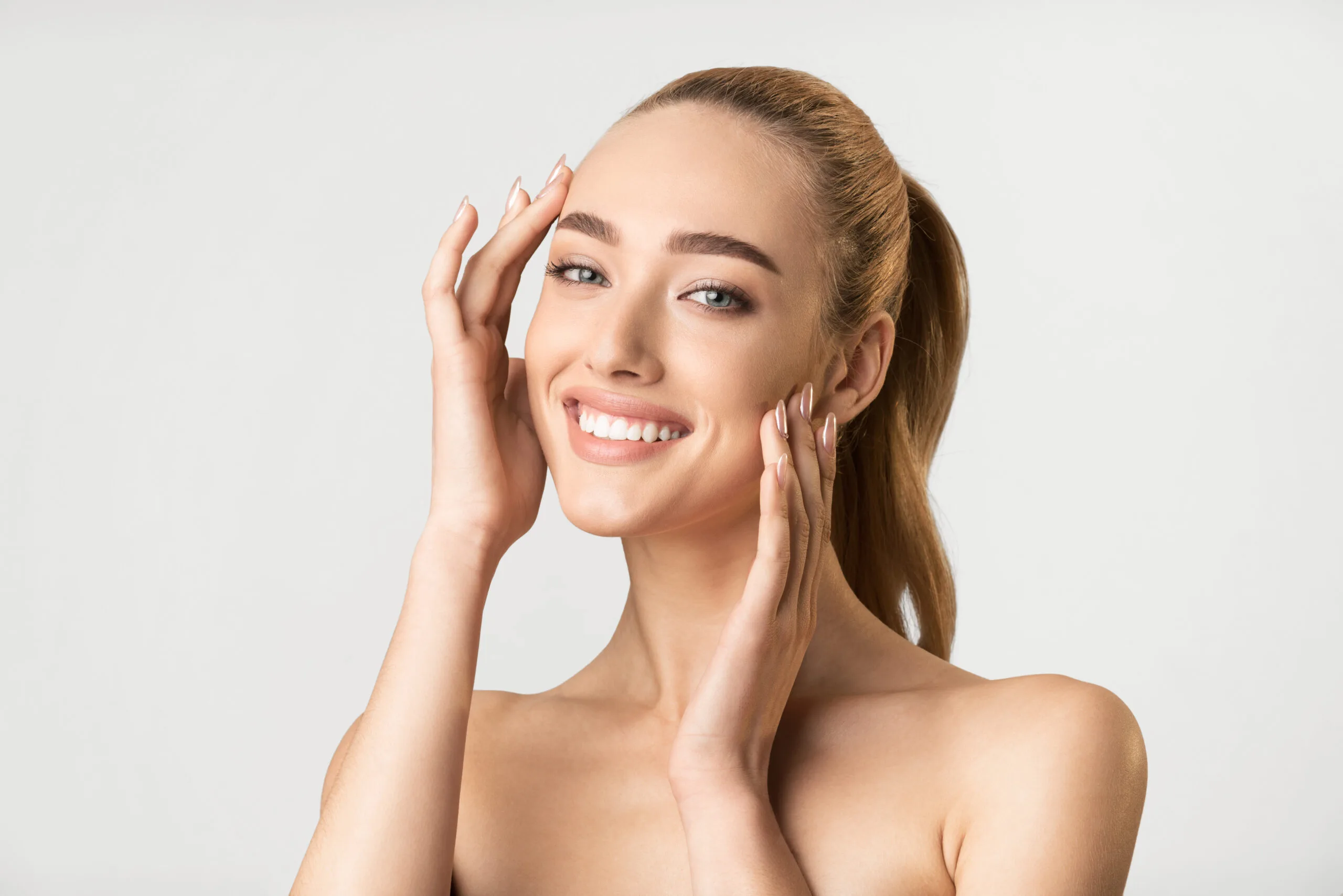 Woman with clear skin smiling and touching her face against a neutral background