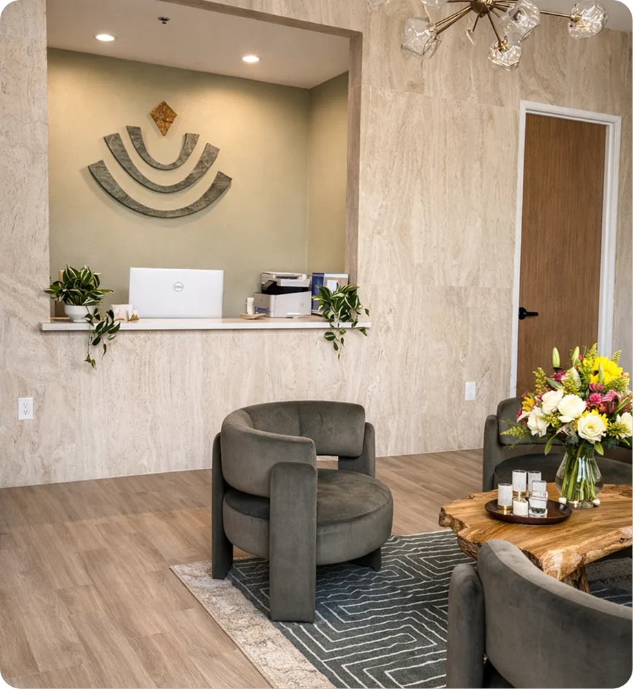 Reception area with curved gray chair, wooden desk with white laptop, decorative smile wall art, fresh flowers in vase, and stone tile walls