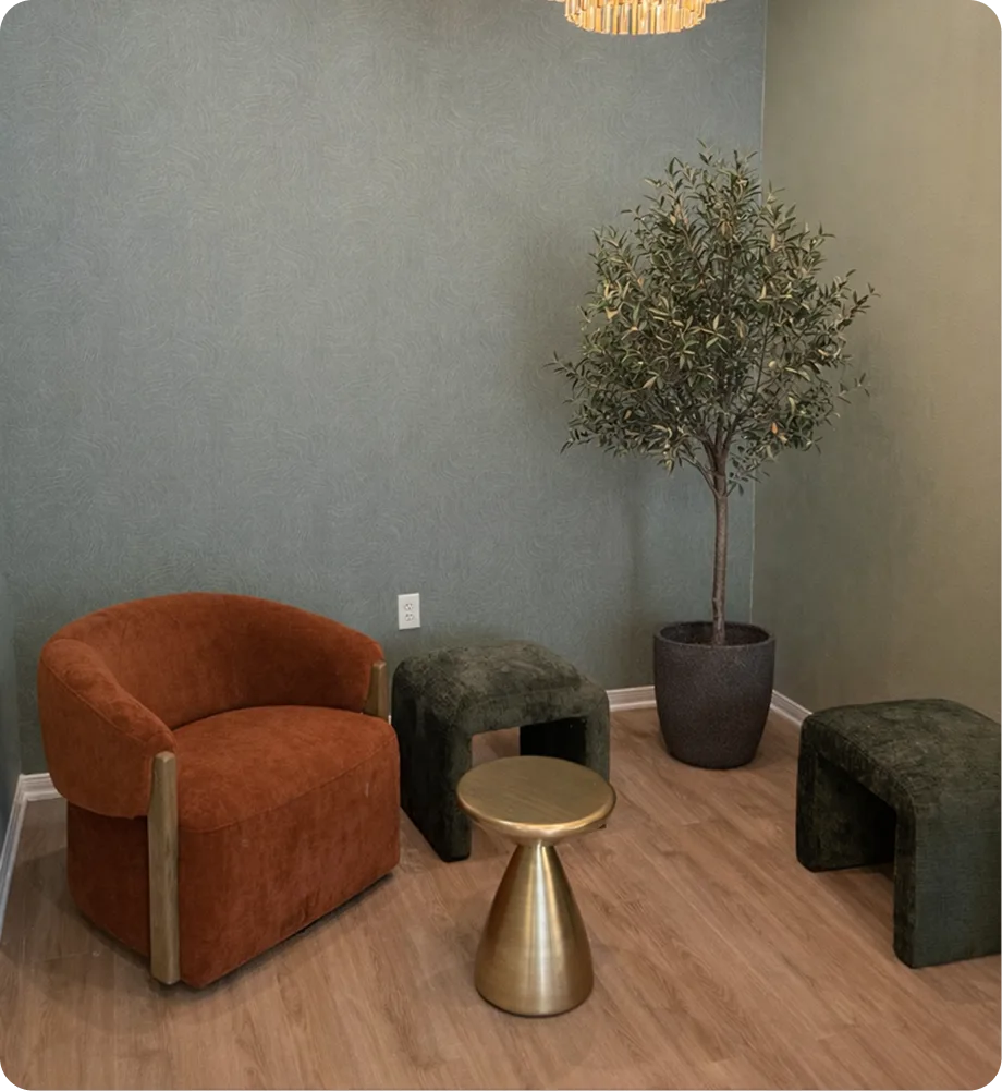 Waiting area with rust-colored upholstered chair, green ottoman seating, brass side table, potted olive tree, and pendant lighting