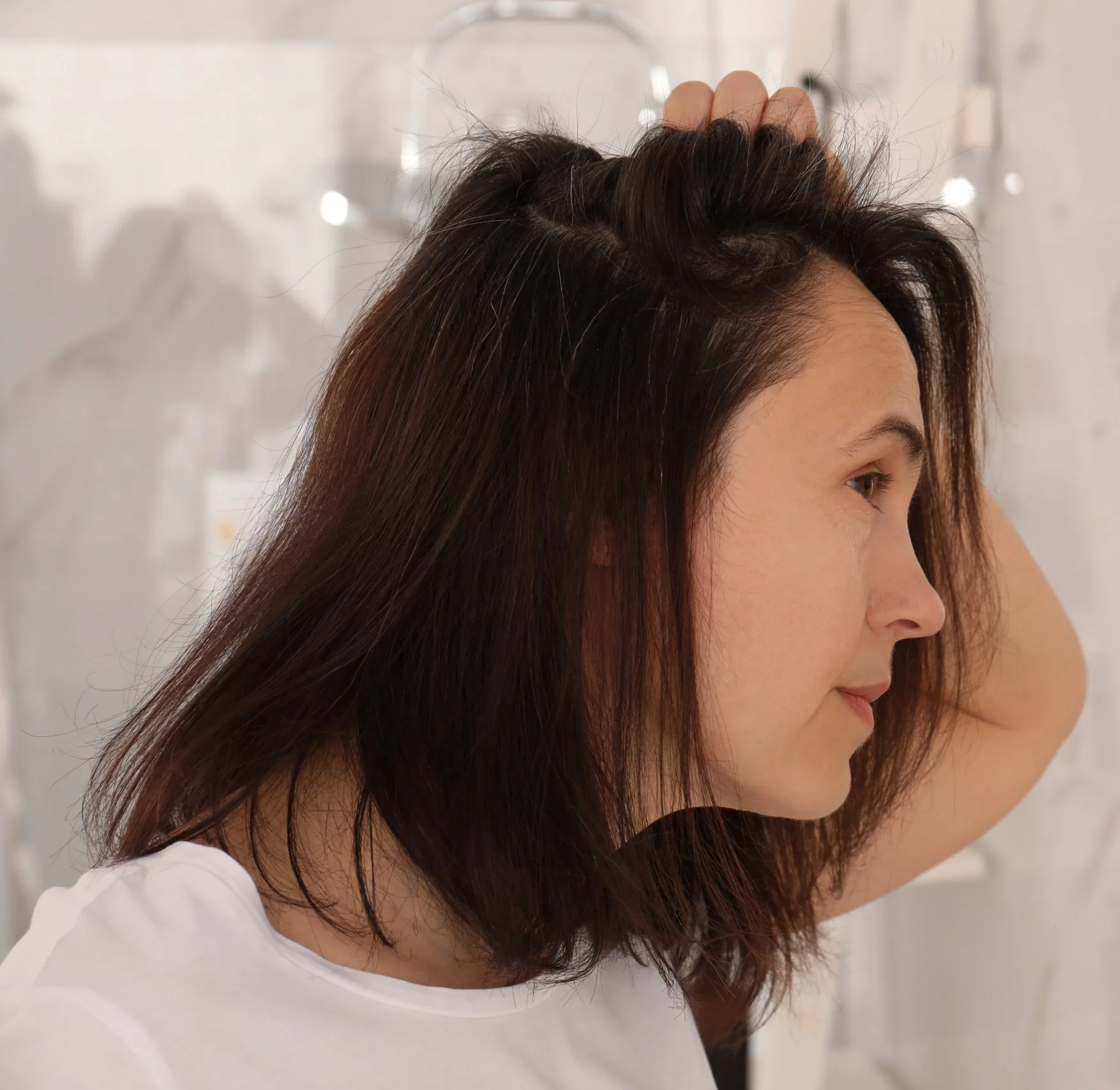 Woman in profile examining her hair and scalp with her hand near bathroom mirror