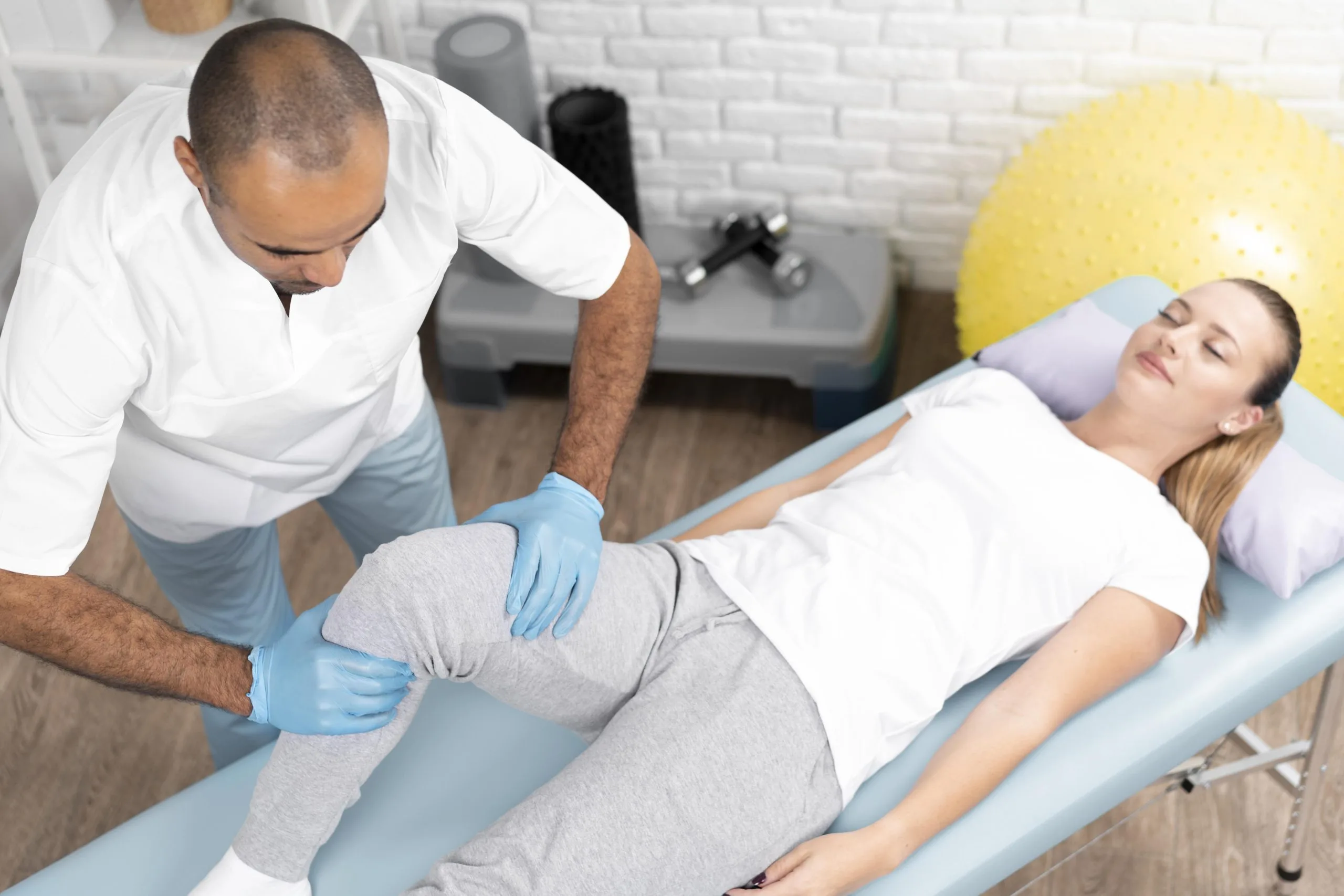 Physiotherapist performing knee assessment on woman lying on treatment table in clinic