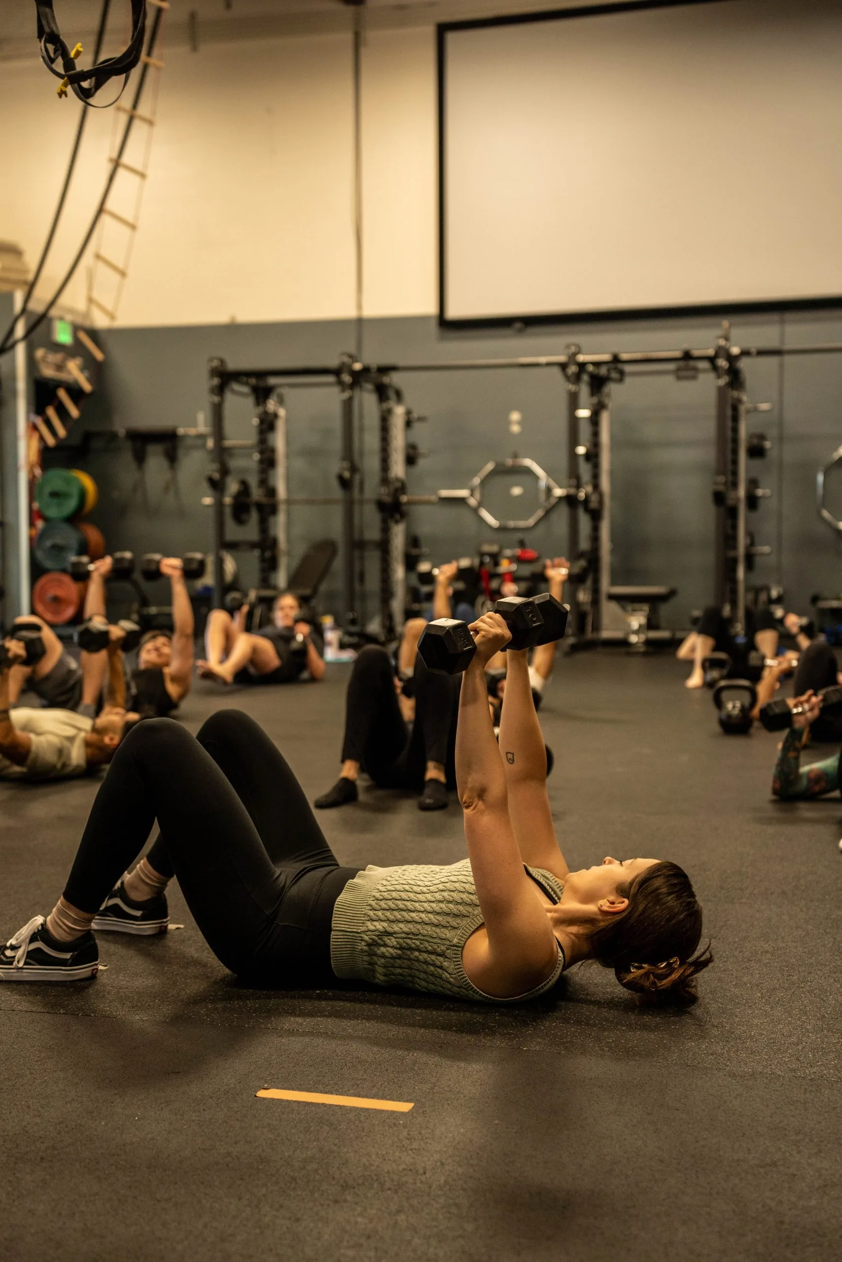 Woman doing dumbbell chest press exercise on gym floor during group fitness class with equipment and participants visible