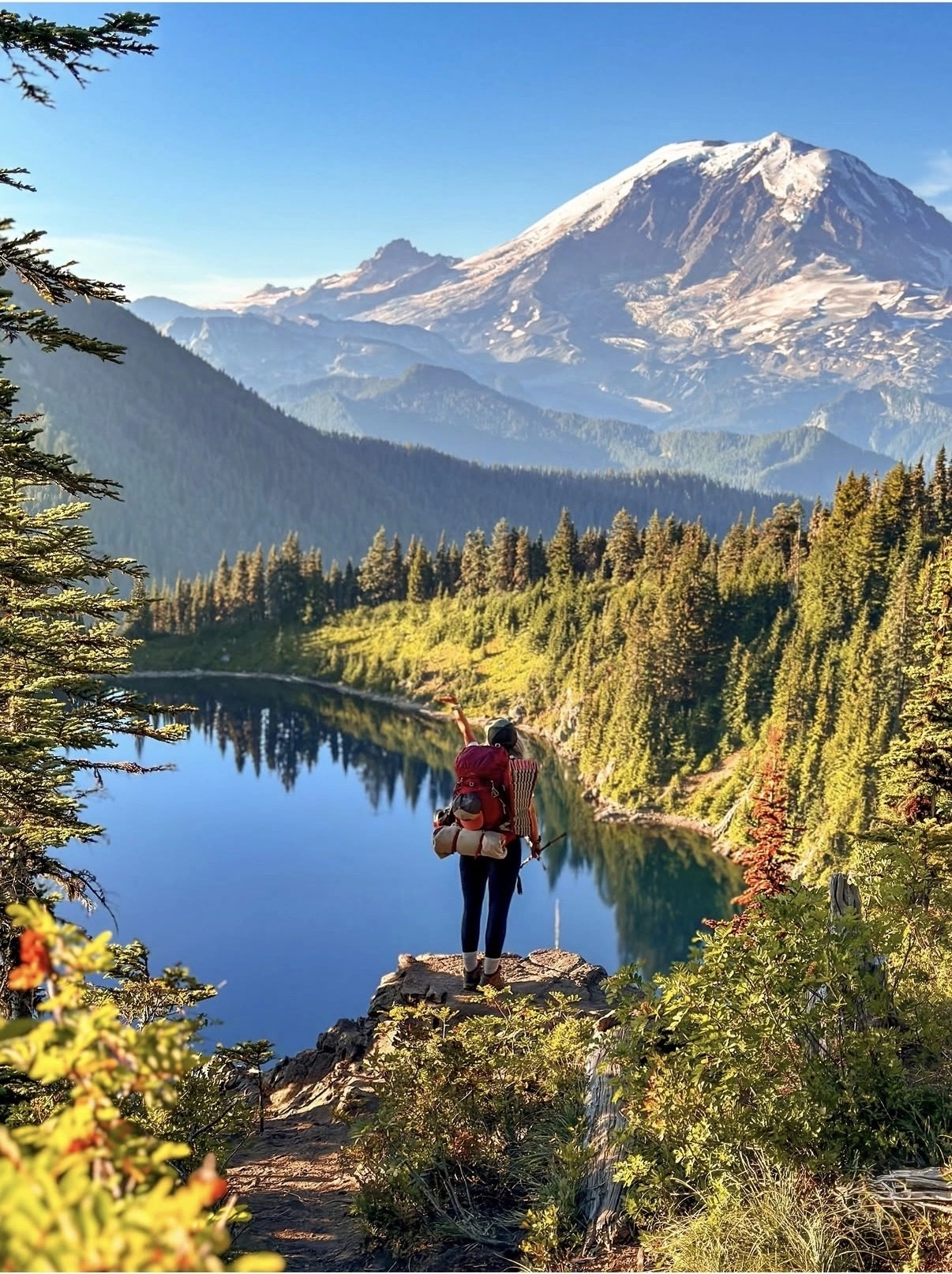 Hiker with red backpack standing on rocky outcrop overlooking alpine lake and snow-capped mountain