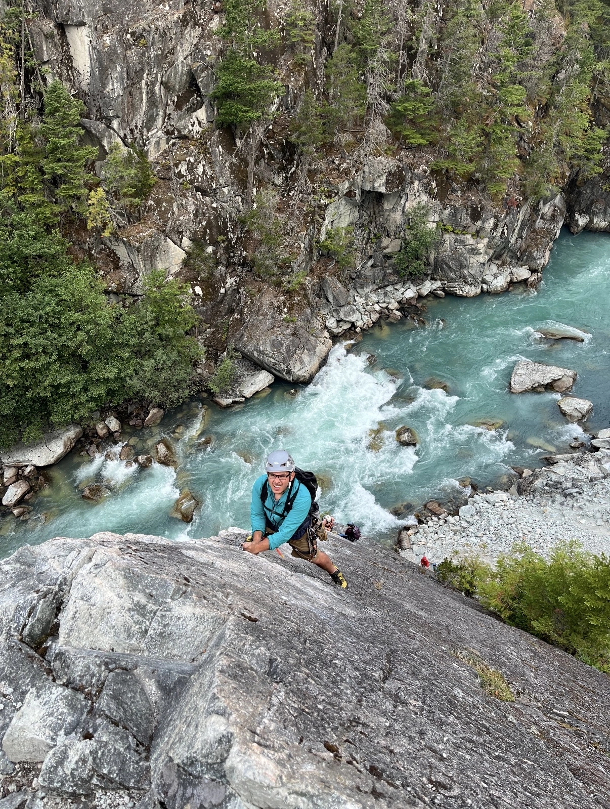 Person rock climbing rappelling down a cliff face above turquoise river with moss-covered rocks and evergreen forest