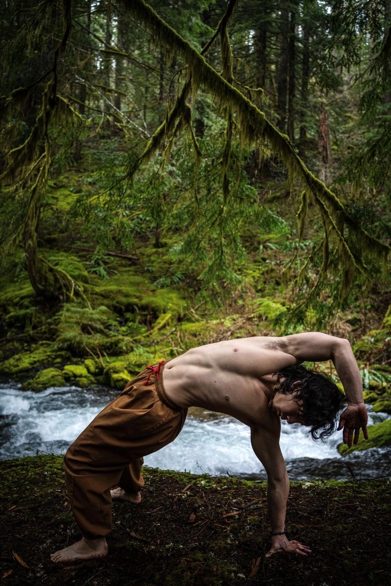 Person performing backbend yoga pose by flowing stream in moss-covered forest with evergreen trees