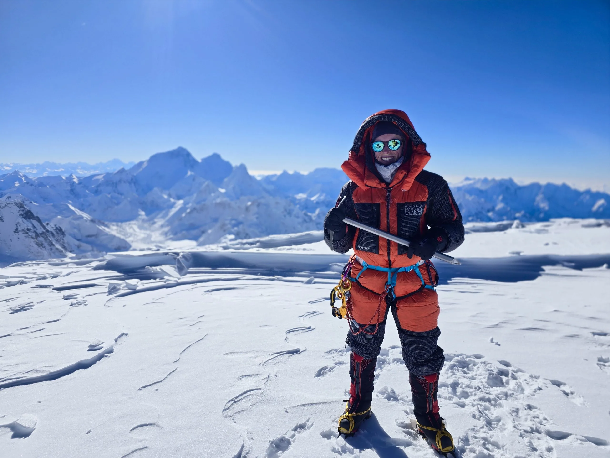 Nicole in red and black mountaineering gear standing on snowy peak with distant mountain range