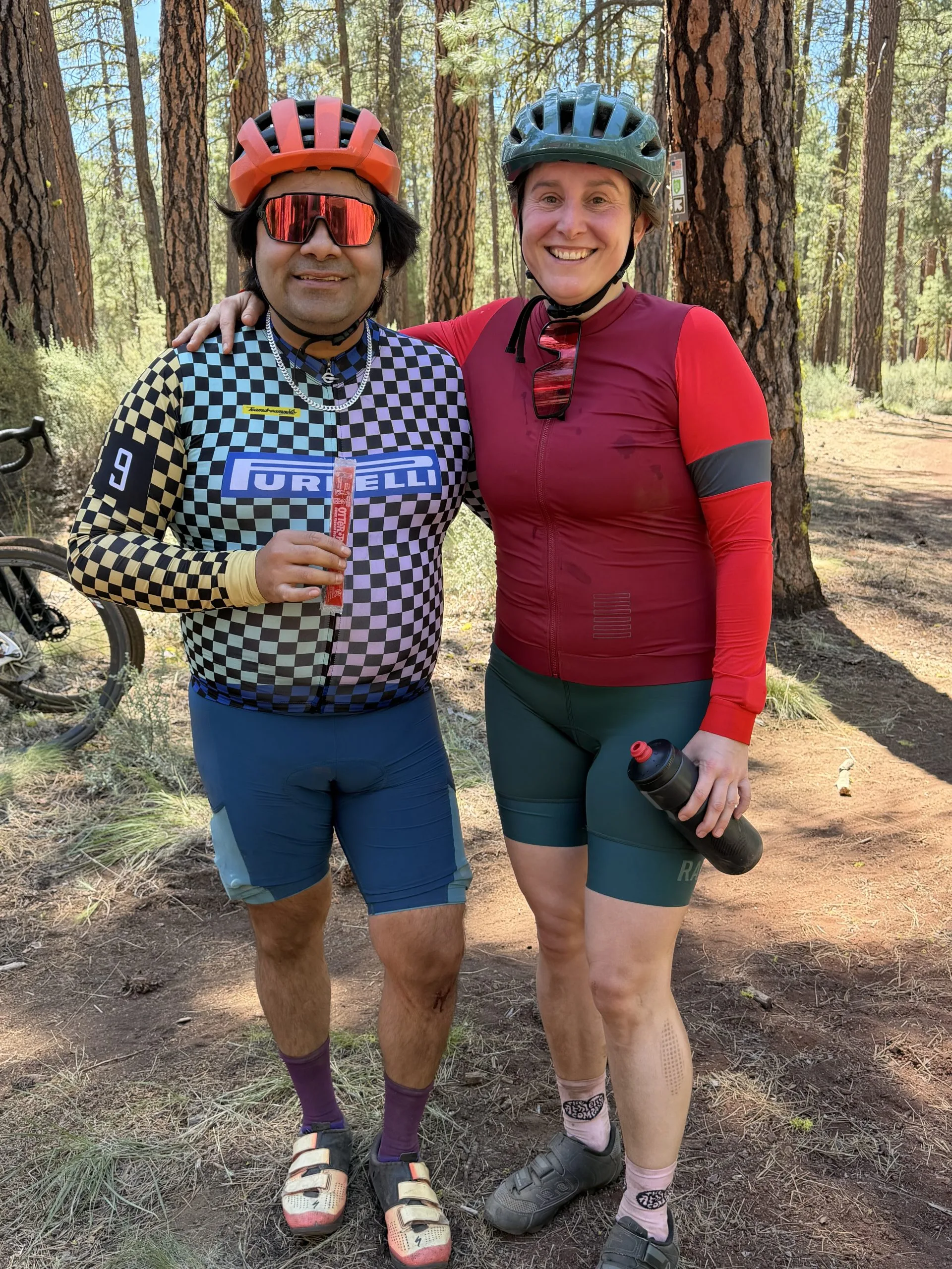 Two cyclists wearing helmets and athletic gear posing together on a forest trail