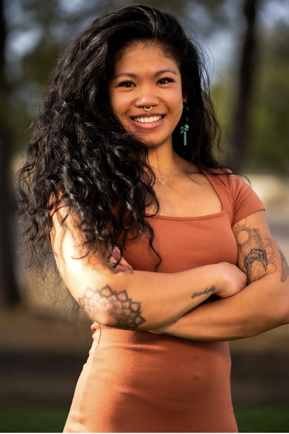 Smiling practitioner with long dark hair and tattoos, wearing rust-colored top, posed outdoors