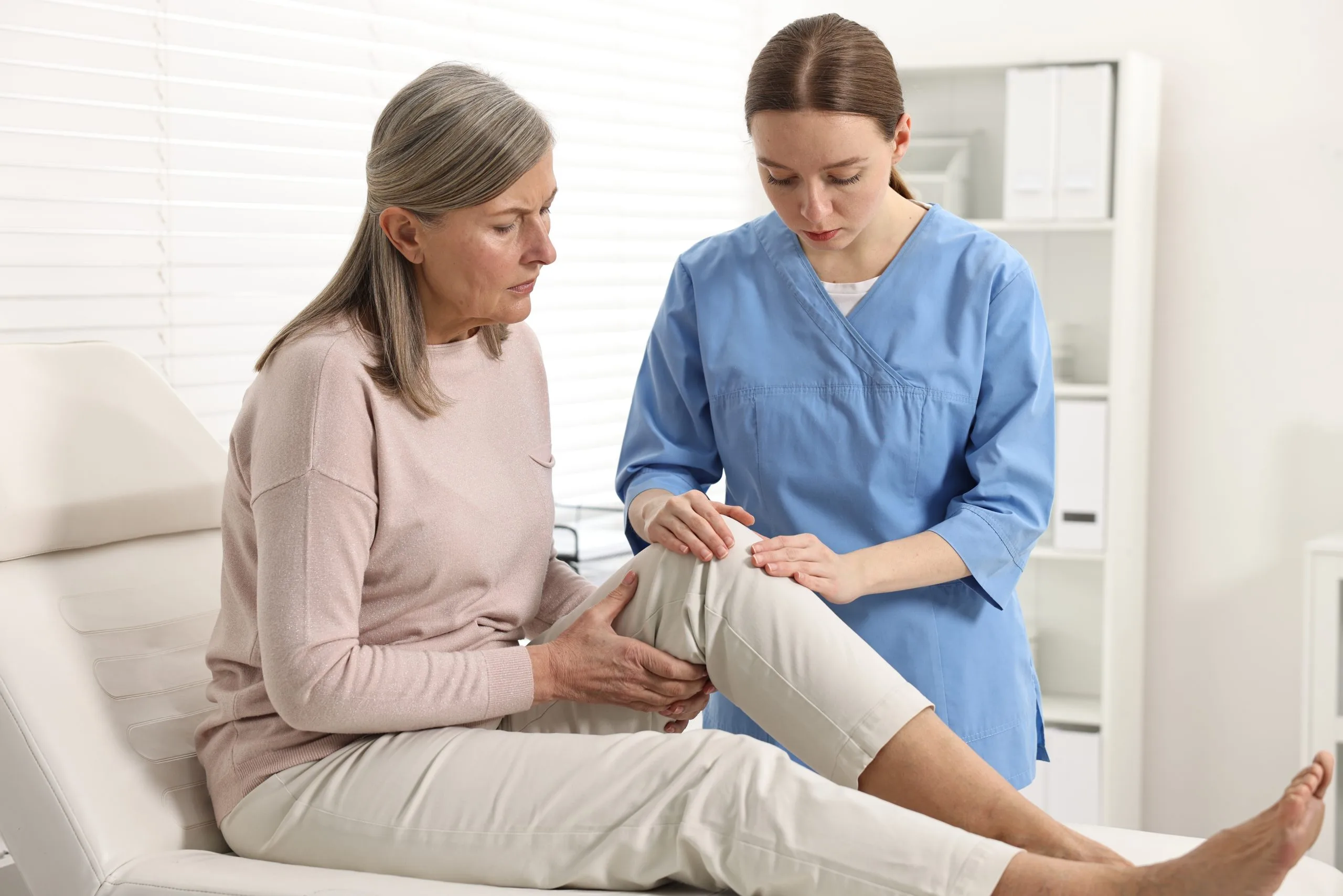 Healthcare provider in blue scrubs examining patient's knee while patient sits on examination table