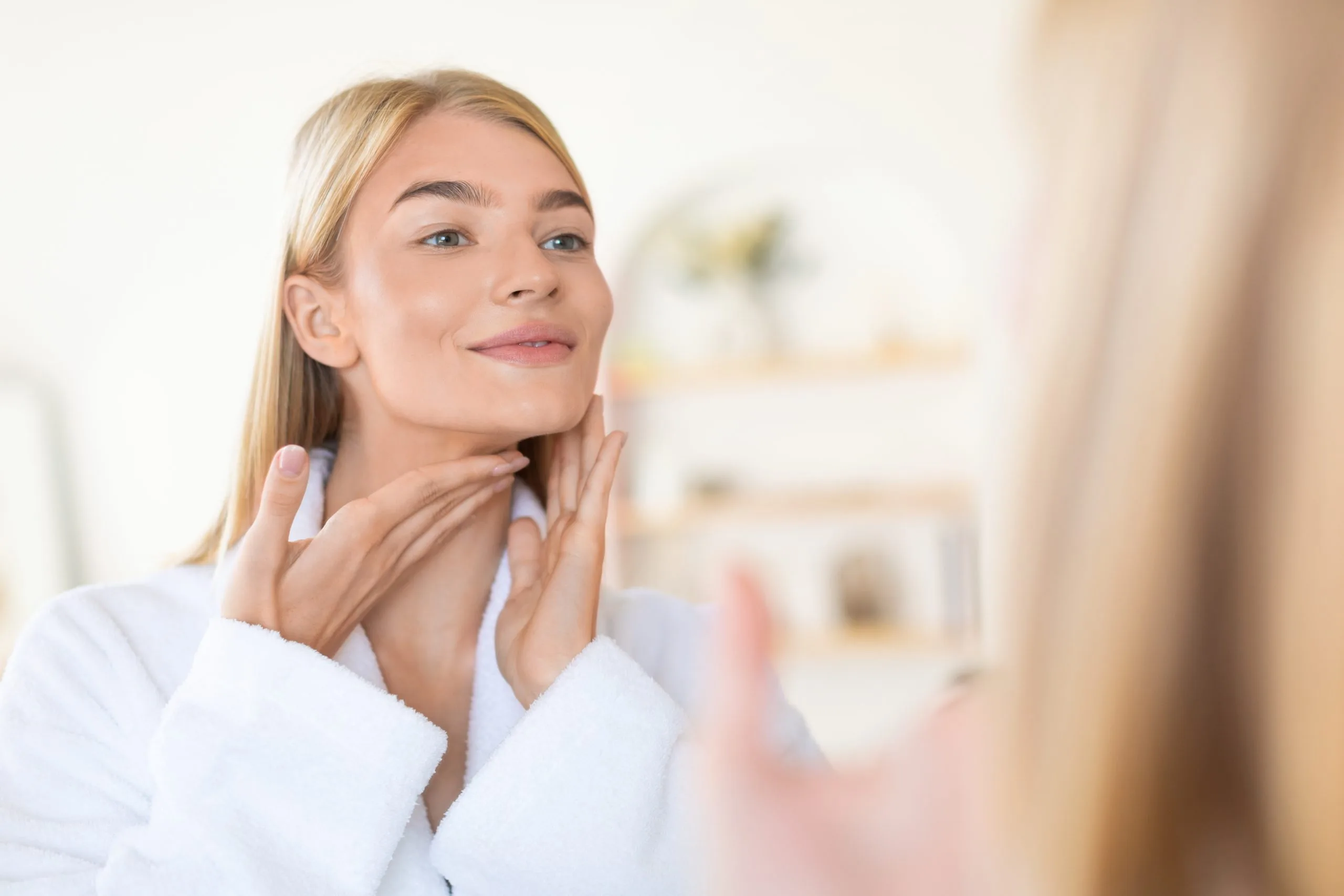 Woman in white shirt touching neck and jaw in bathroom mirror