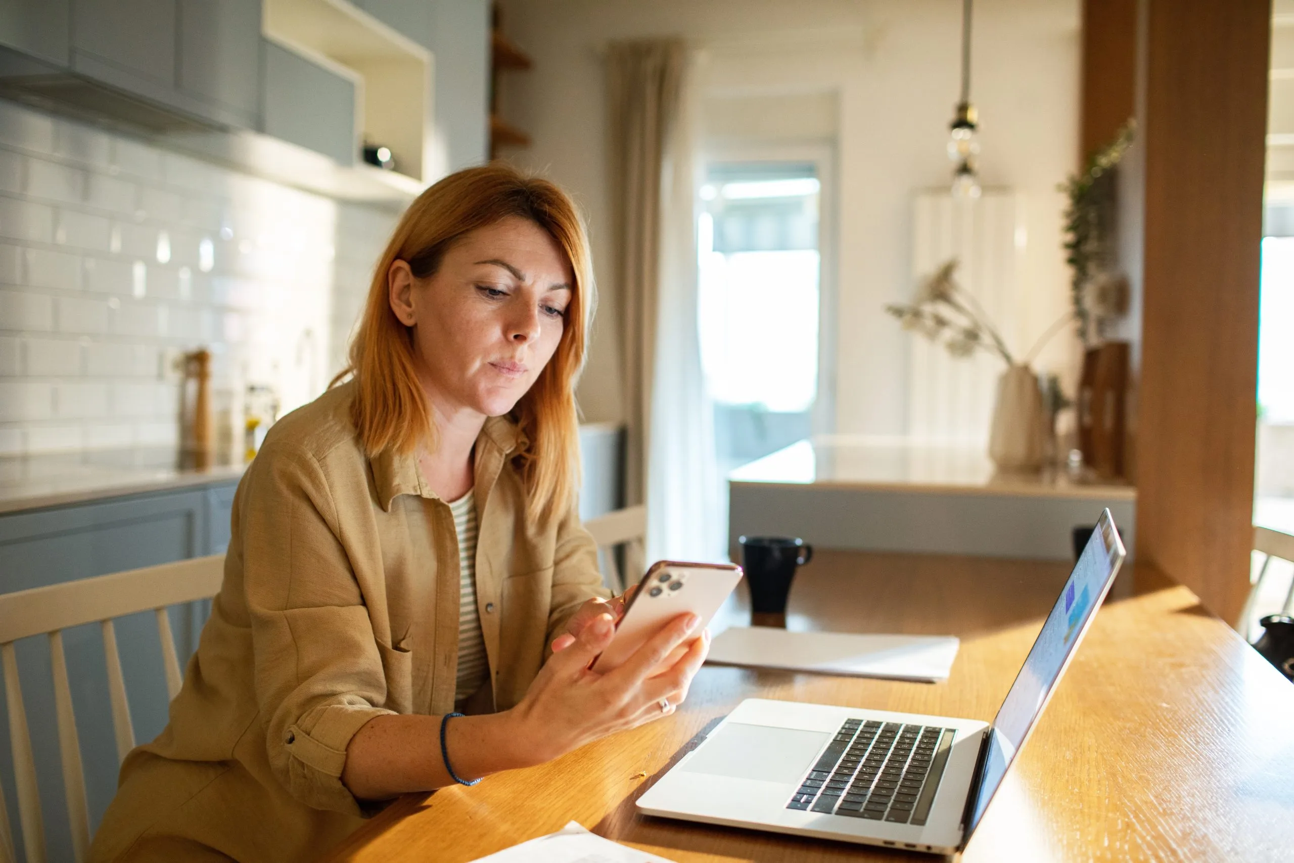 Woman with red hair holding a smartphone at a wooden table with a laptop and coffee cup