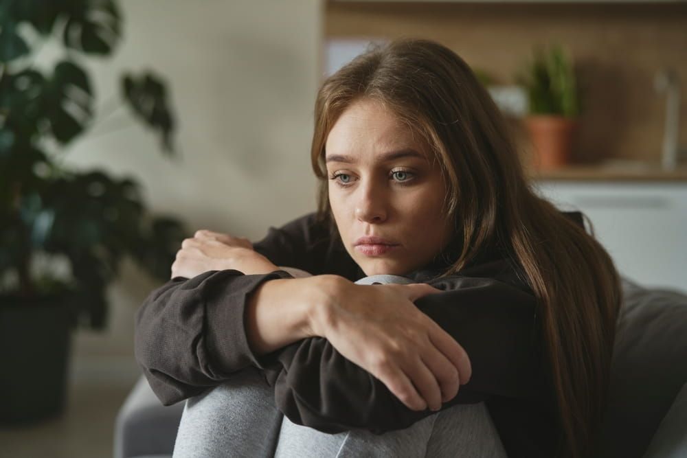 Woman with downturned expression hugging her knees while seated, suggesting emotional distress or fatigue