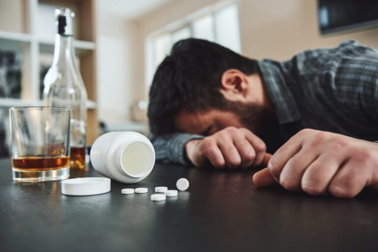 Man slumped over table with alcohol bottle, glass, and scattered pills