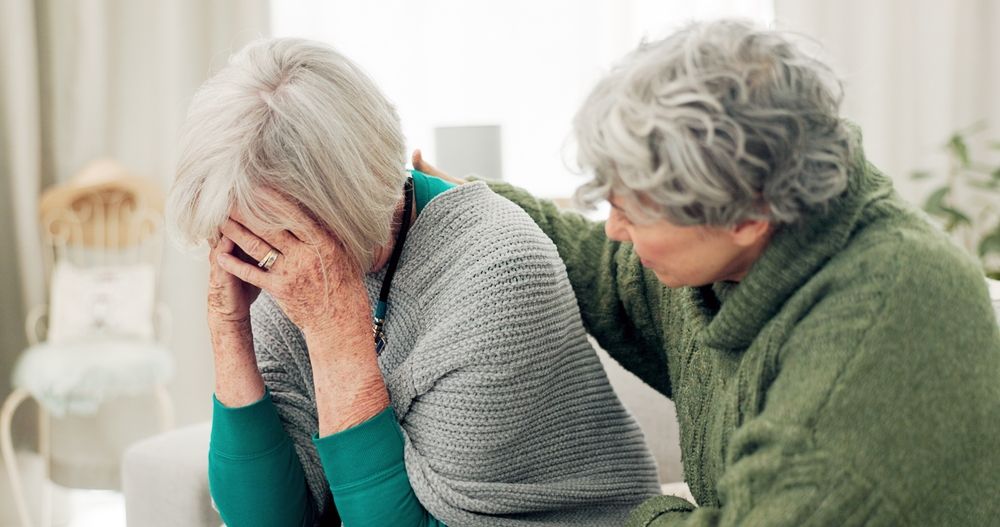 Older woman holding her head in distress while another woman comforts her