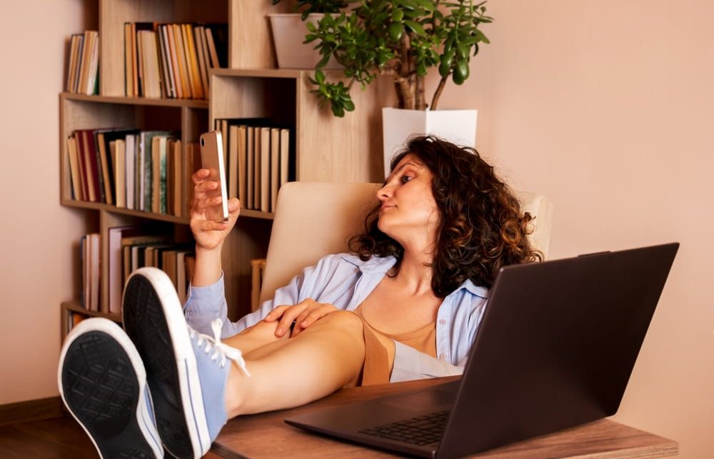 Woman relaxing at desk with feet up, holding a book while working on laptop