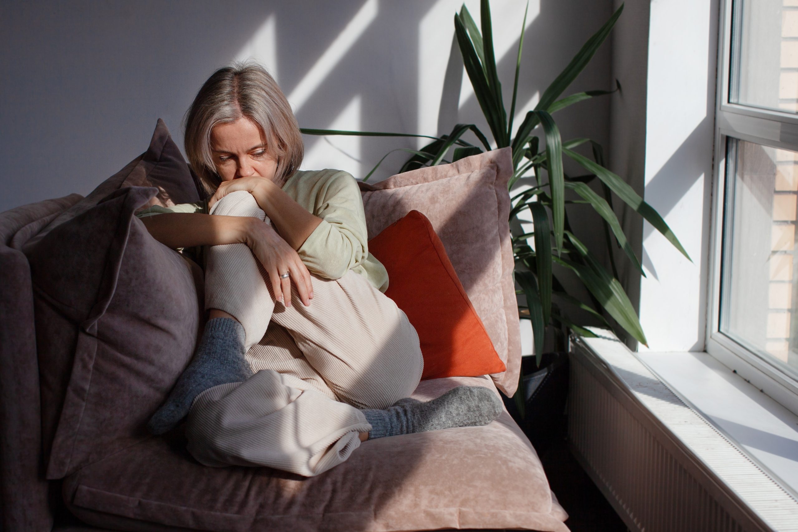 Woman sitting on couch near window with potted plant, hand to face in contemplative pose