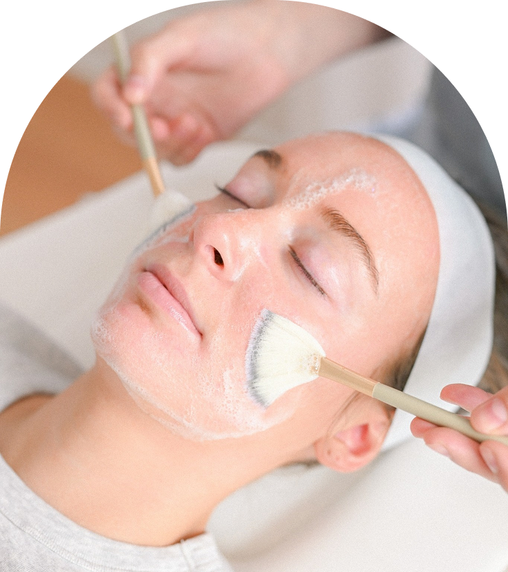 Aesthetician applying white facial treatment mask to relaxed client's face with brush