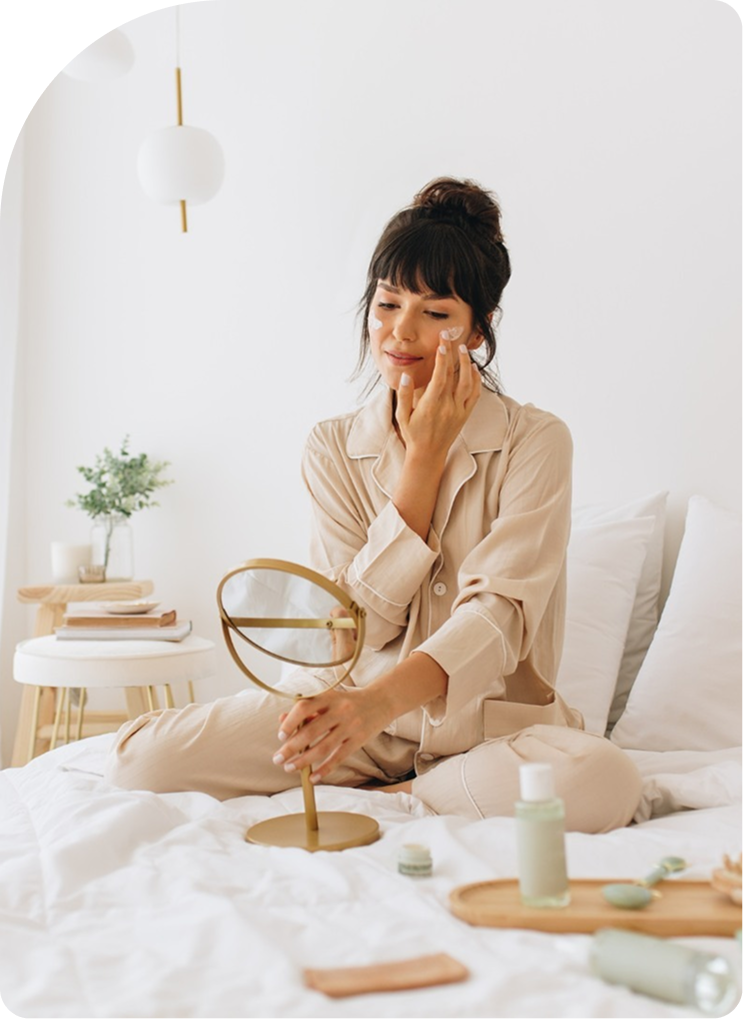 Woman applying skincare product to her face while sitting on a bed with aesthetic products displayed
