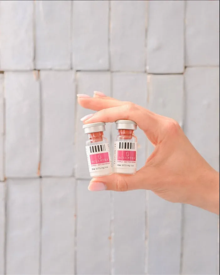 Two vials of Sculptra injectable treatment held in hand against white tile backdrop