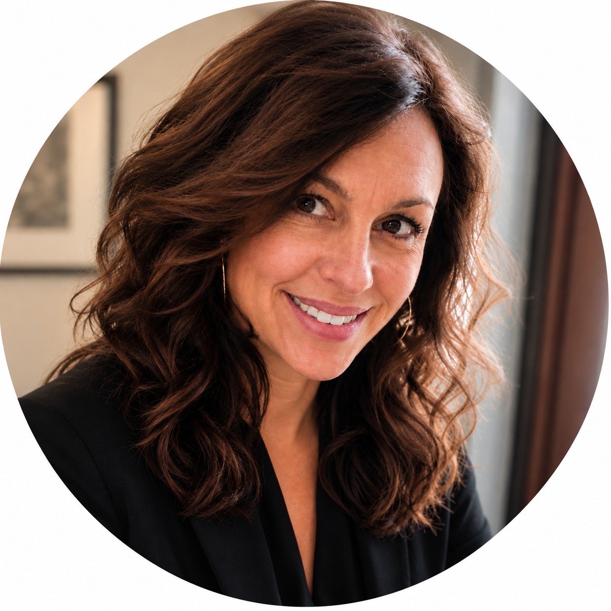 Woman with brown wavy hair smiling at camera, wearing black blazer and hoop earrings