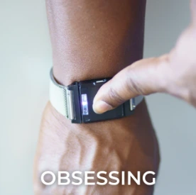 Person's arm wearing a dark smartwatch with text reading 'OBSESSING'
