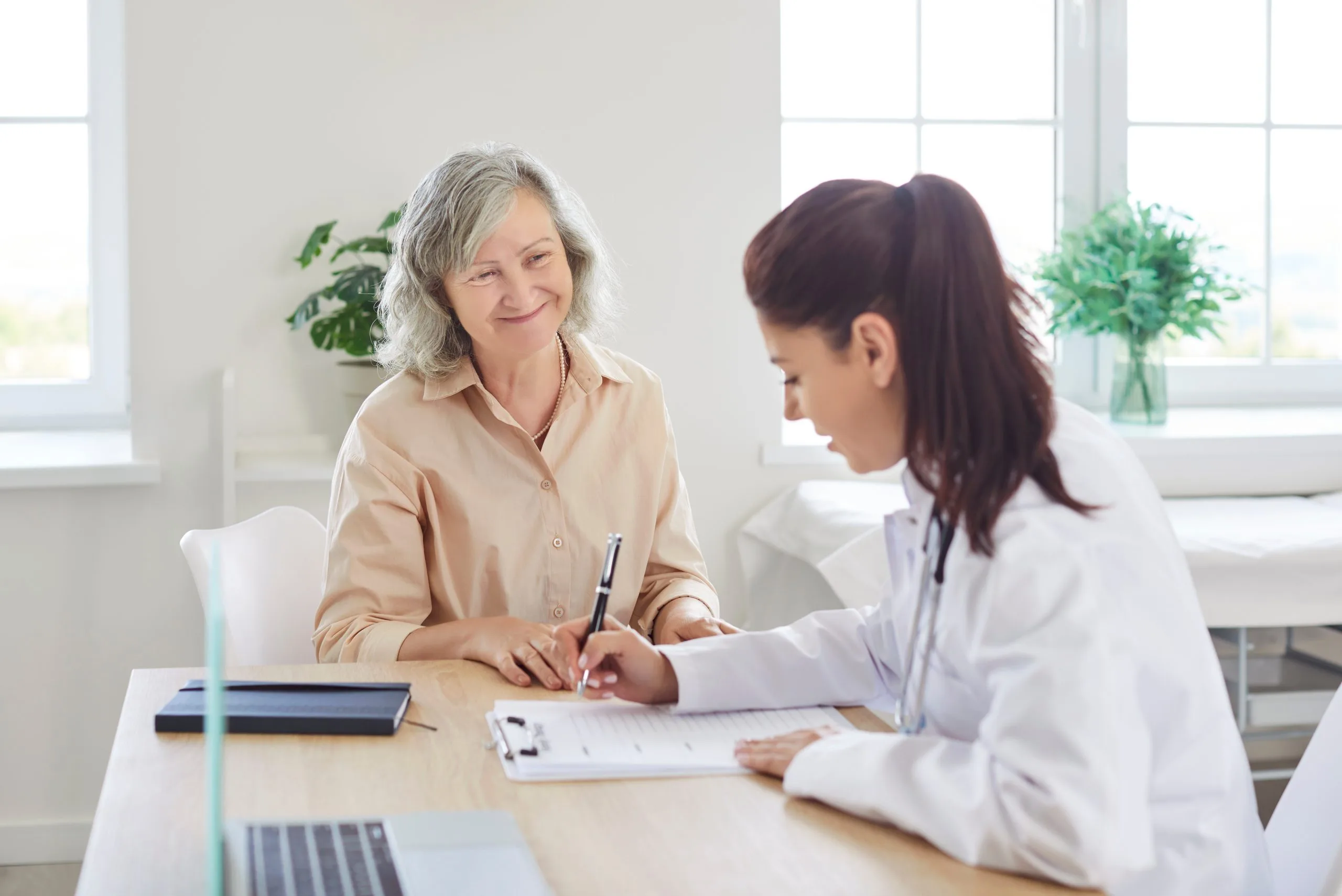 Healthcare provider in white coat and patient signing paperwork at desk in bright office with plants