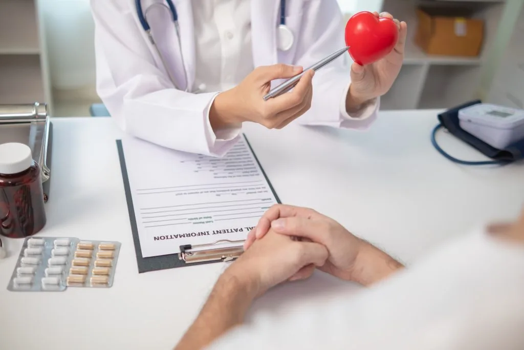 Cardiologist holding red heart model while discussing cardiac care with patient reviewing medical documentation