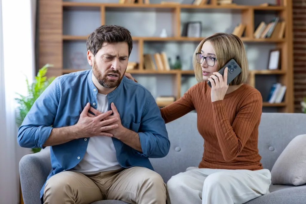 Woman on phone comforting man experiencing chest pain on couch at home