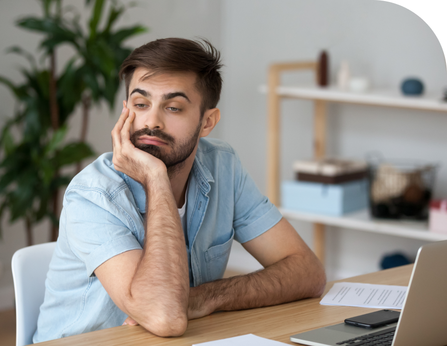 Man sitting at desk with laptop and paperwork, resting chin on hand with concerned expression