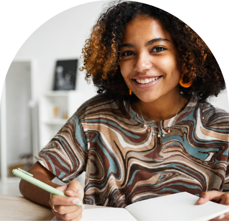 Smiling young person with curly hair holding a pen while writing at a desk