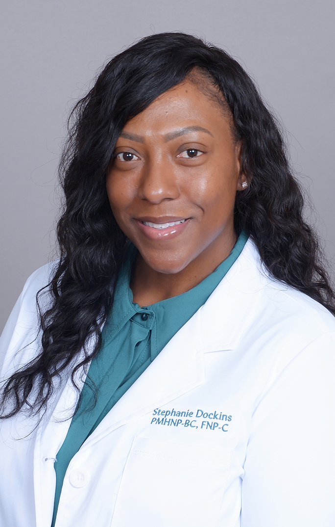 Stephanie Dockins, PMHNP-BC, FNP-C in white coat with teal shirt against gray background