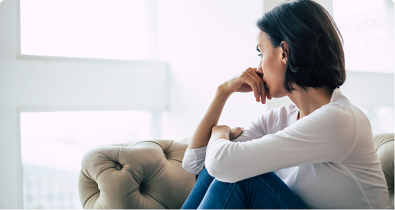Woman in white shirt sitting on couch with thoughtful expression, hand to chin