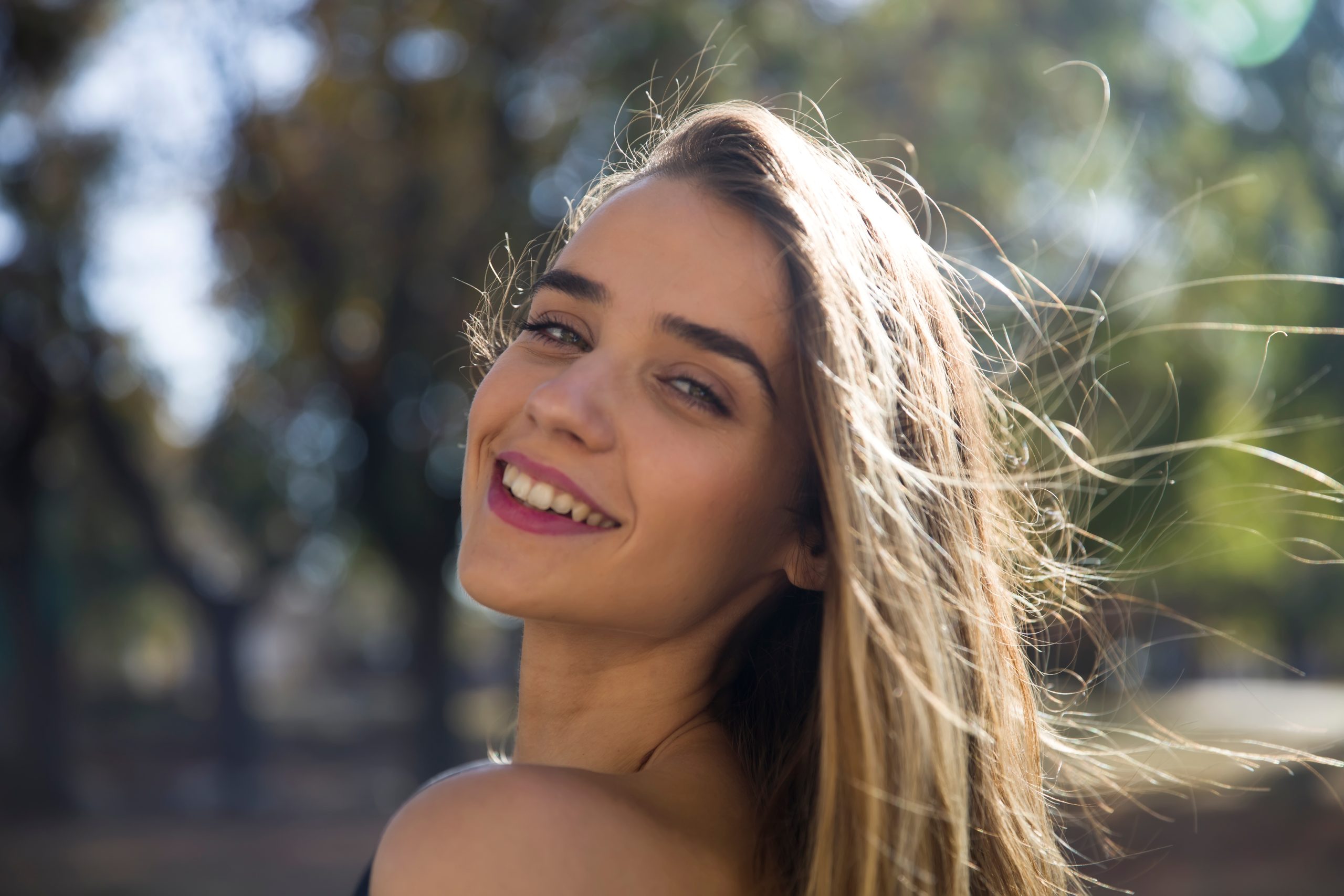 Woman with long hair smiling in natural sunlight near trees