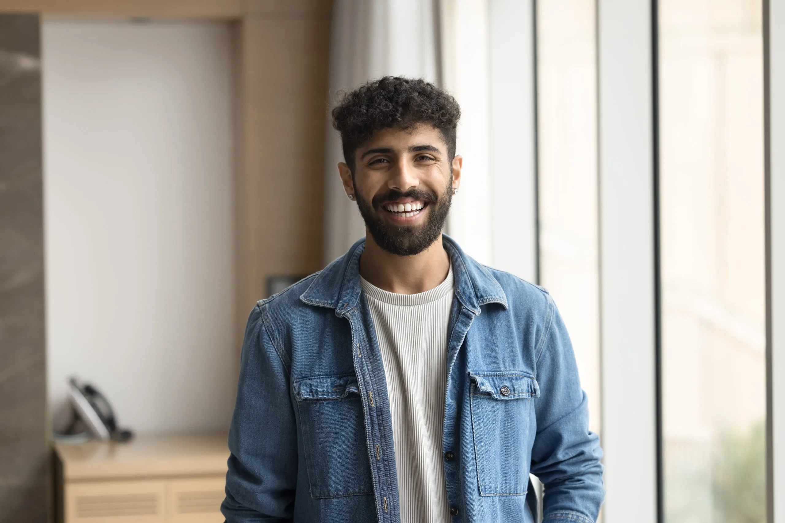 Man smiling in modern hallway wearing denim shirt over white ribbed top