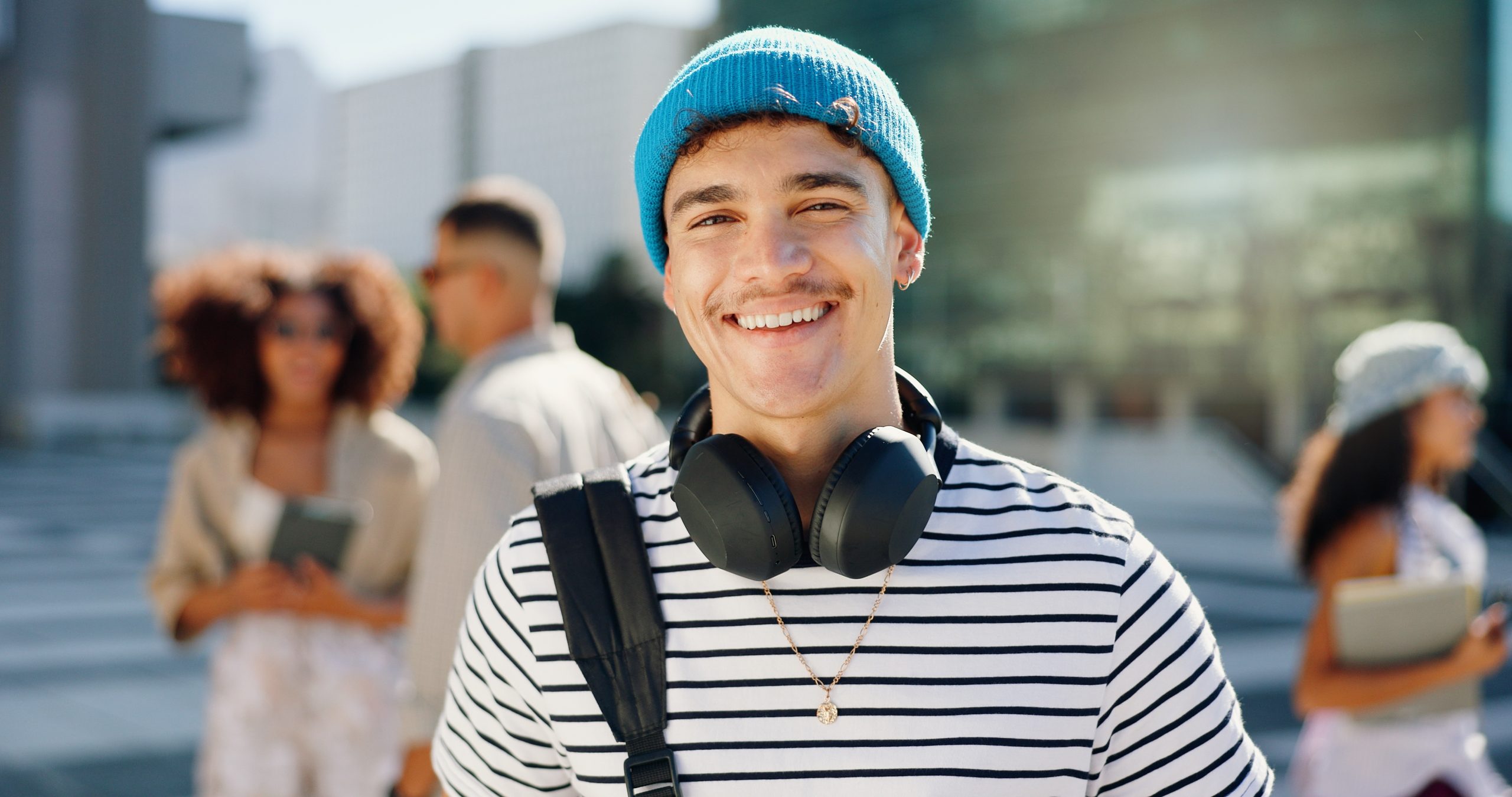 Young man smiling outdoors wearing blue beanie and headphones with backpack