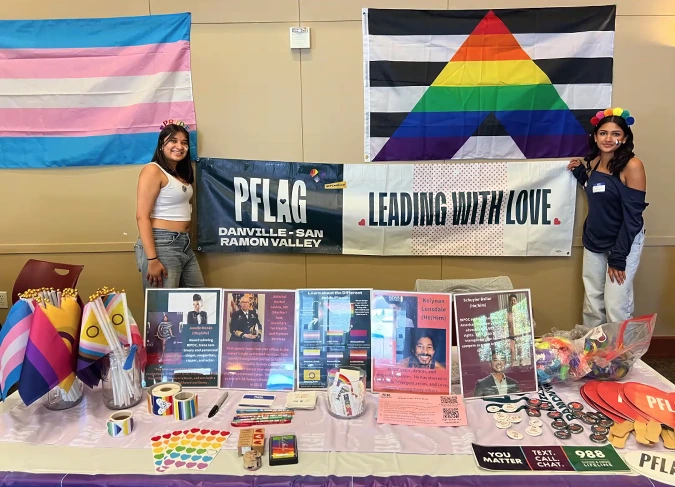 Two people holding PFLAG Danville-San Ramon Valley and Leading With Love banners at community table with pride flags and educational materials
