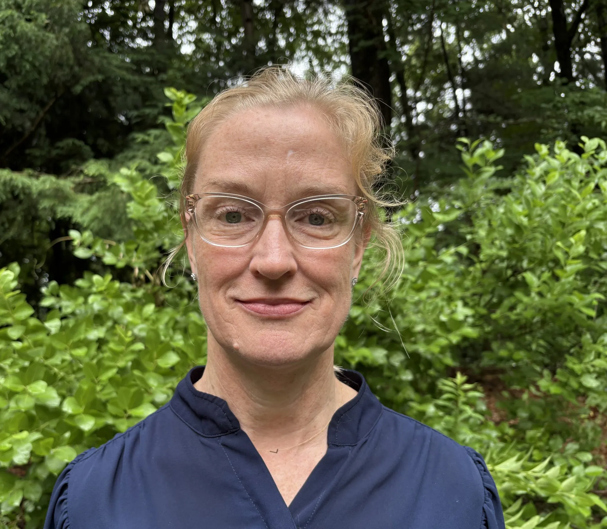 Andrea Butterfield, woman wearing glasses and dark navy shirt outdoors with green foliage background