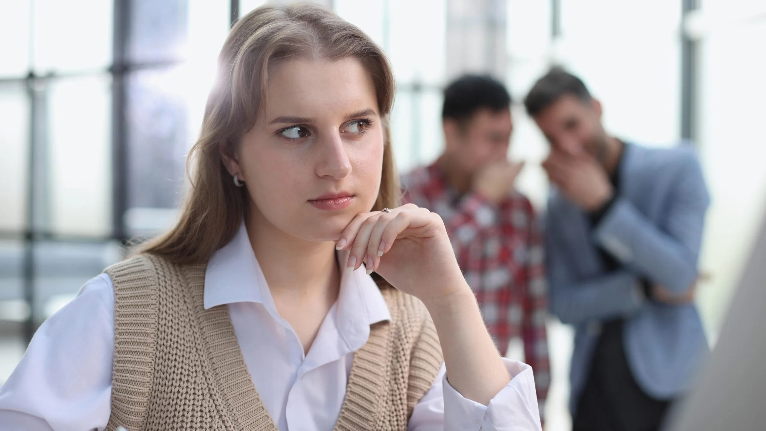 Young woman looking troubled with hand to face while two people converse in background