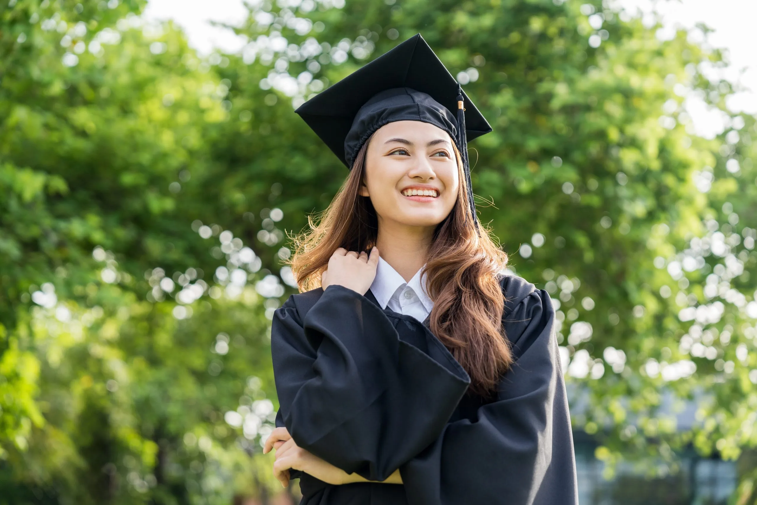 Woman in graduation cap and gown smiling outdoors with green trees in background
