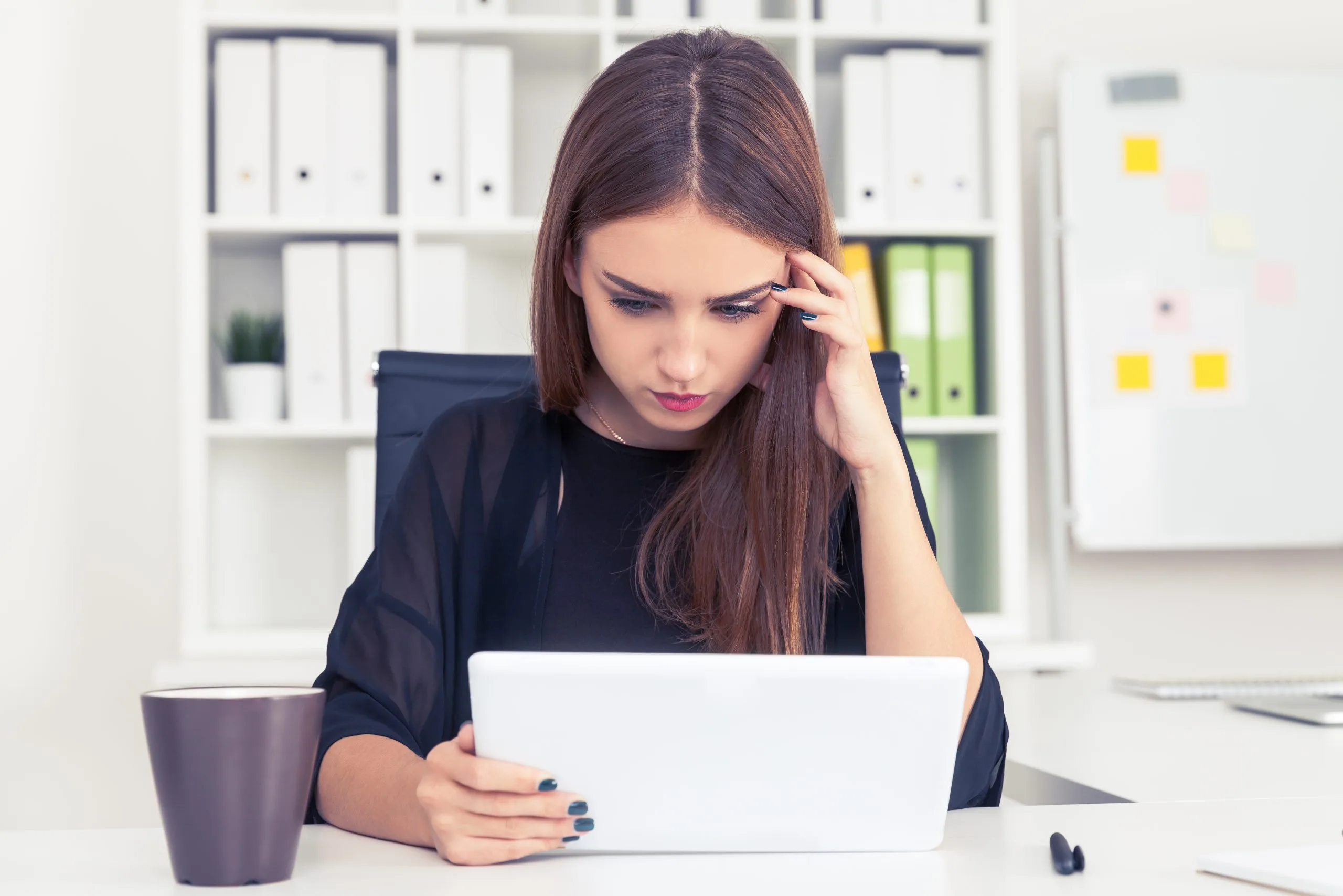 Woman in dark shirt looking at laptop while touching her head at desk with coffee cup and yellow sticky notes