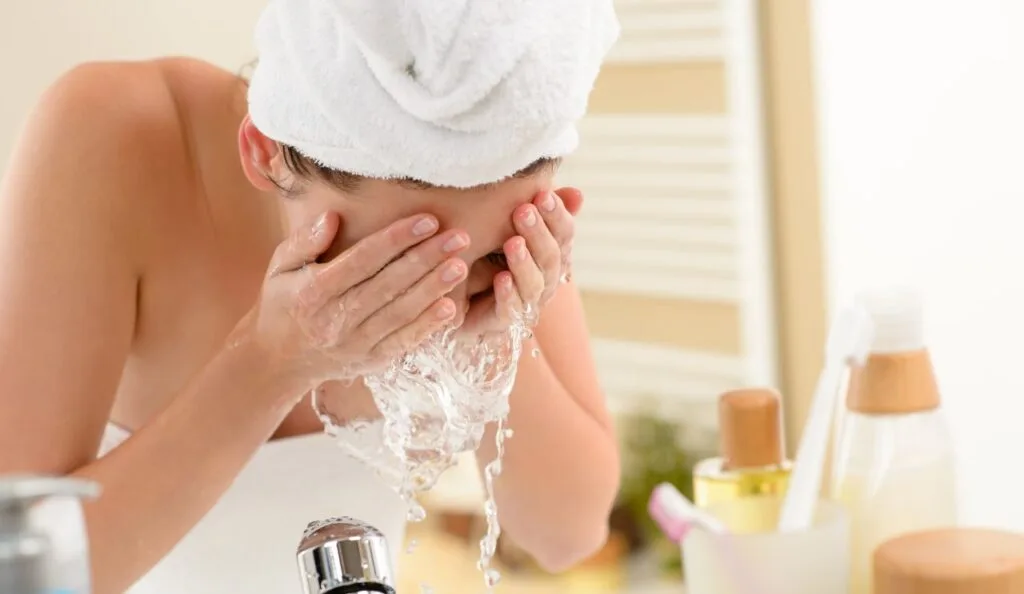 Person with towel-wrapped head rinsing face with water over a sink