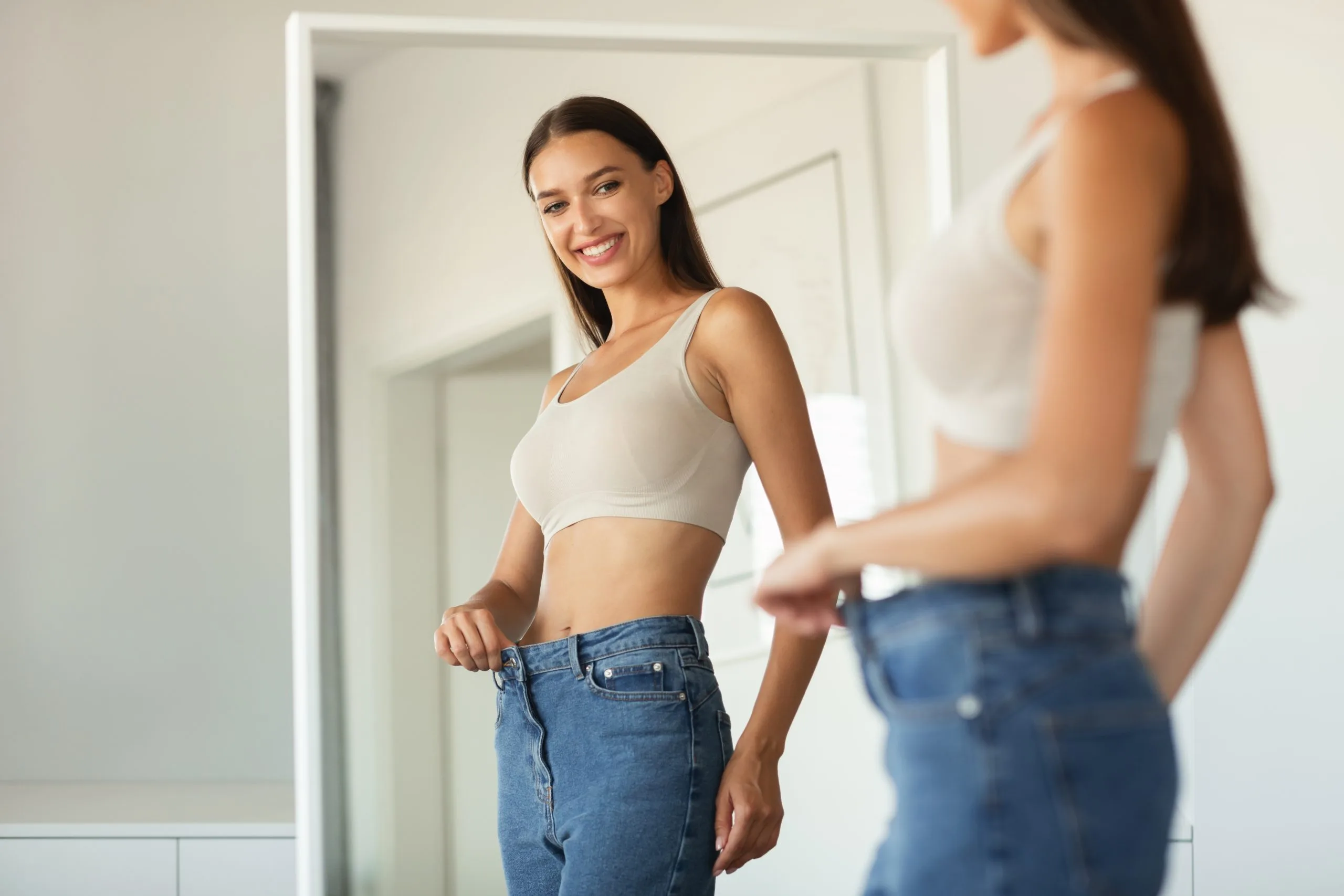Woman holding loose jeans at waist, smiling at mirror reflection after weight loss