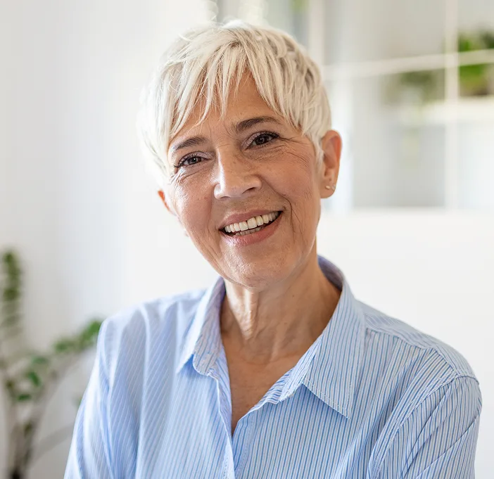 Smiling woman with short white hair wearing a light blue striped shirt in a bright clinical setting