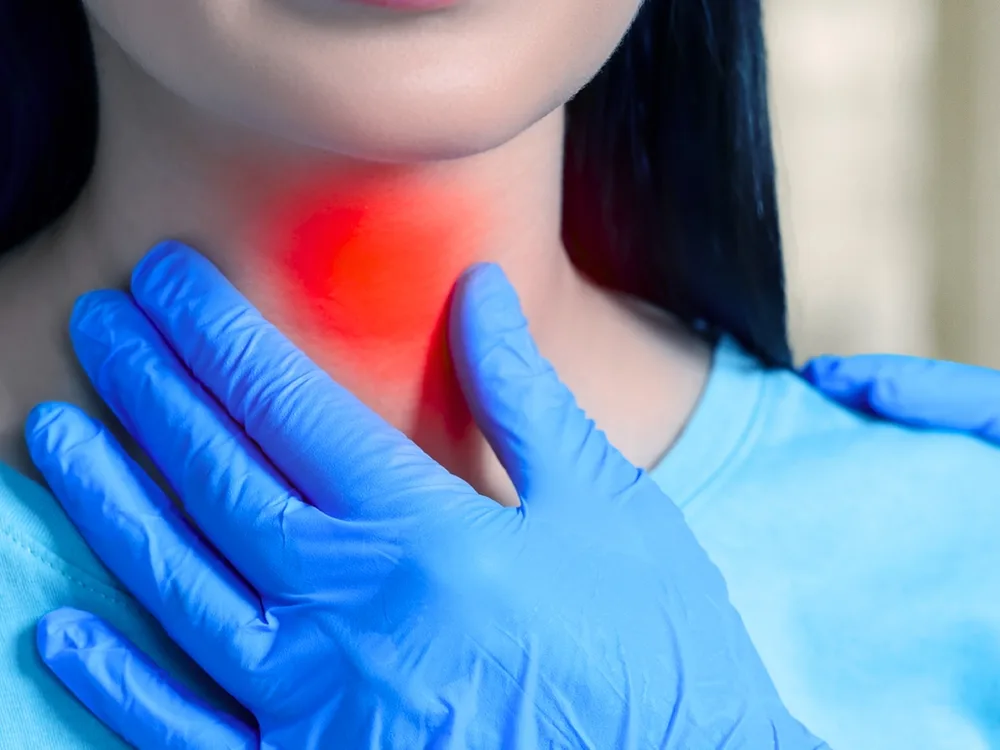 Woman in blue gloves pointing to red inflamed area on her neck, indicating thyroid pain