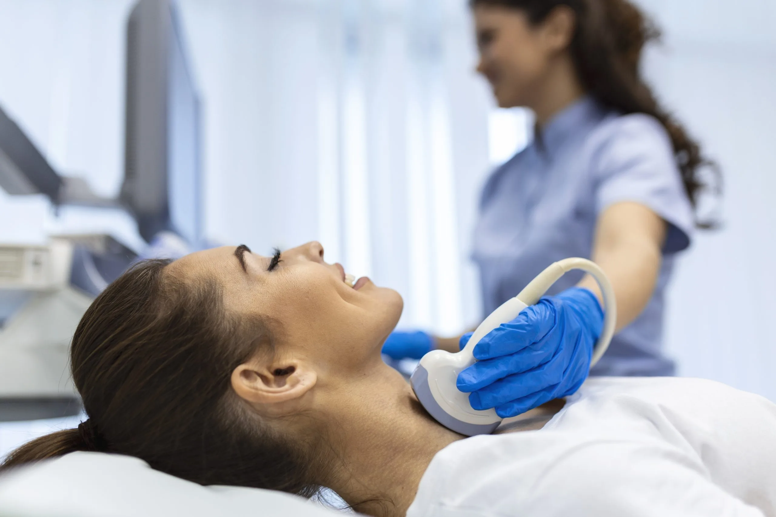 Clinician performing thyroid ultrasound on woman's neck with probe while doctor observes in background