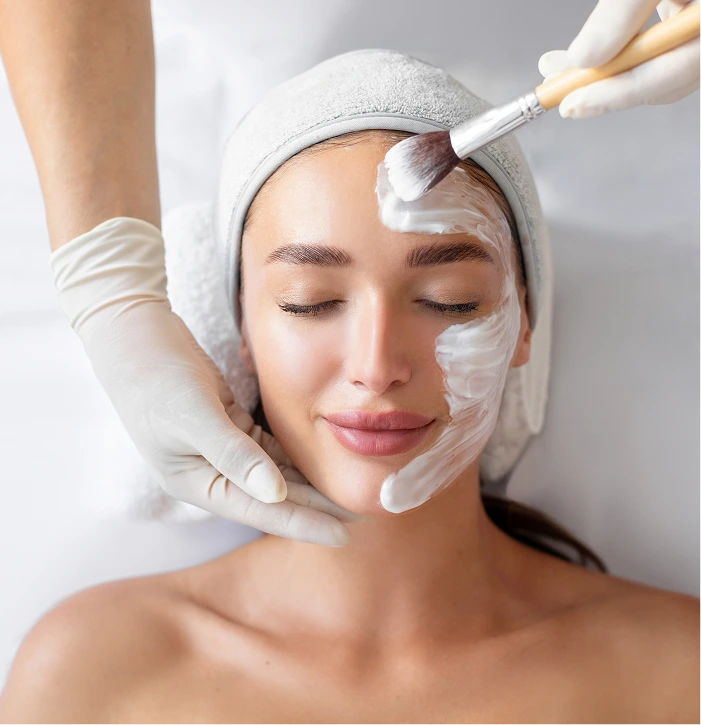 Esthetician applying treatment to woman's face with brush while wearing head wrap and gloves