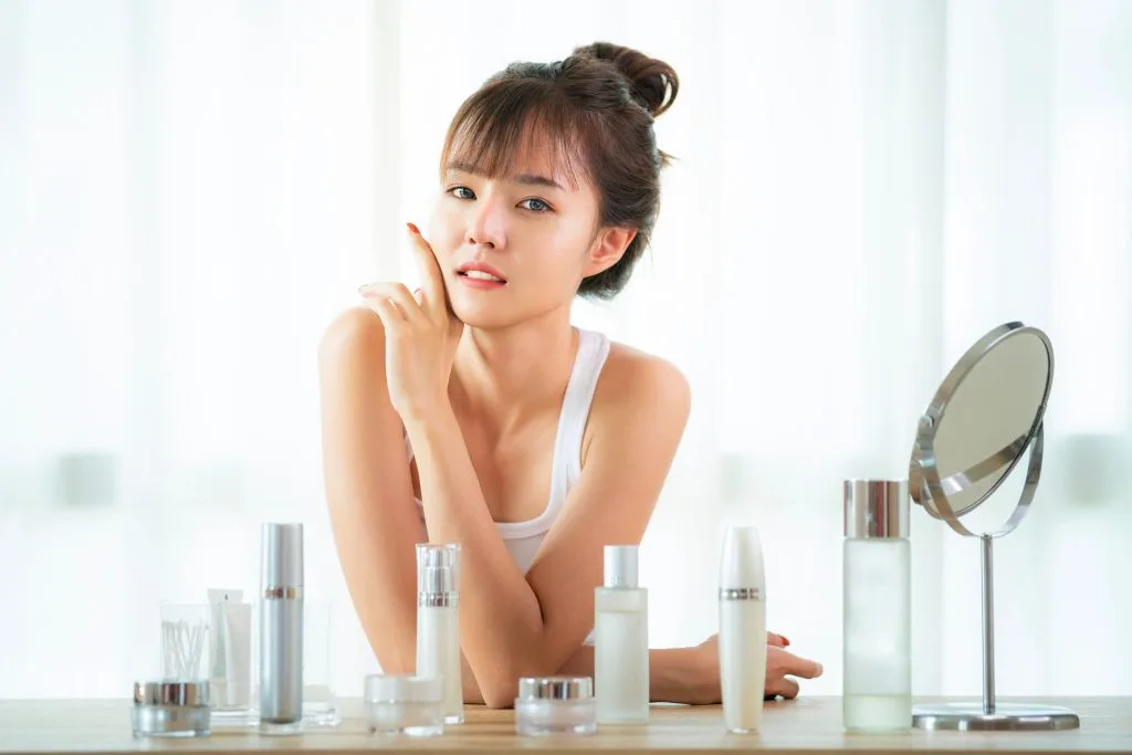 Woman examining her face in handheld mirror surrounded by skincare product bottles