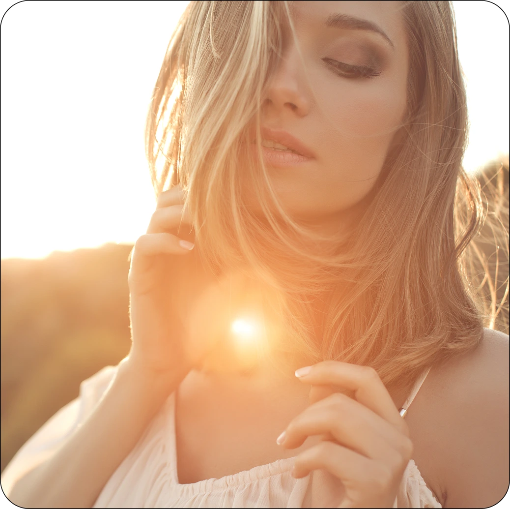 Woman with blonde hair in golden sunlight, touching her shoulder with a relaxed expression